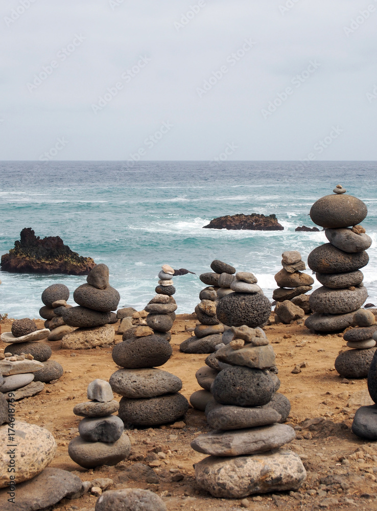 piles of stacked stones and pebbles in zen towers on a yellow sandy beach with sea breaking over rocks and blue calm sky piles of stacked stones and pebbles in zen towers on a yellow sandy beach with sea breaking over rocks and blue calm sky