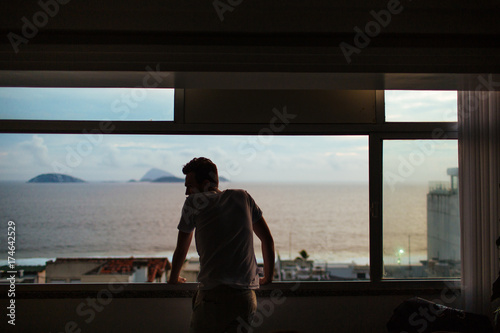 Young man staring out the window onto the beach at sunset 