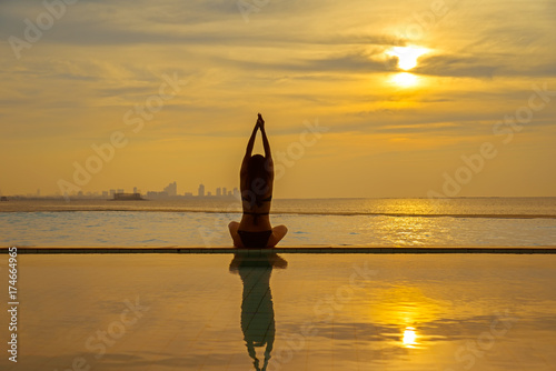 Silhouette young woman practicing yoga on swimming pool and the beach at sunset. Healthy Concept..