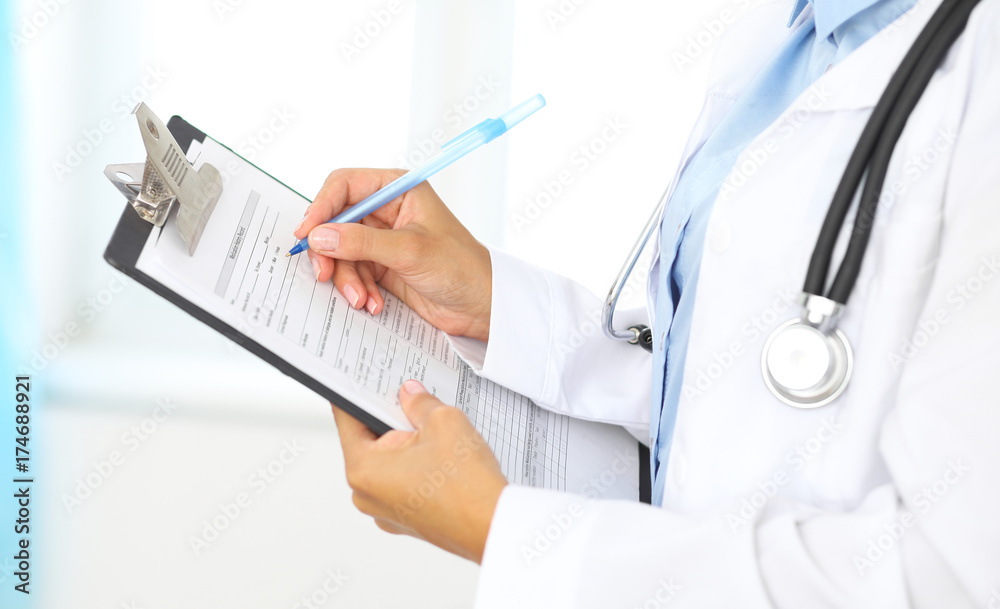 Close-up of a female doctor filling up medical form at clipboard while standing straight in hospital Close-up of a female doctor filling up medical form at clipboard while standing straight in hospital