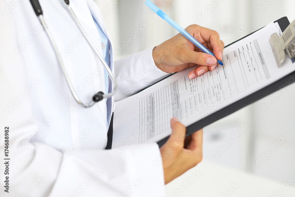 Close-up of a female doctor filling up medical form at clipboard while standing straight in hospital Close-up of a female doctor filling up medical form at clipboard while standing straight in hospital