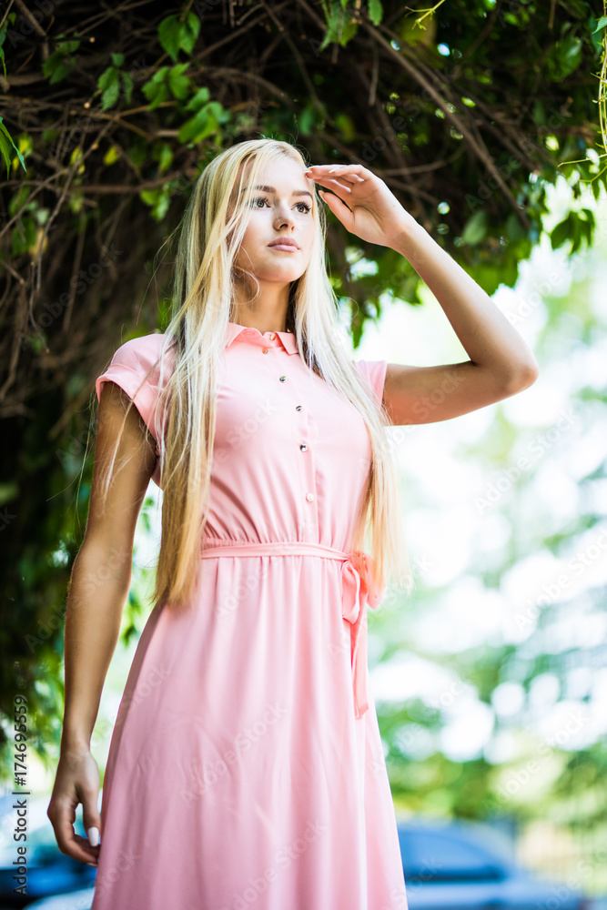 Beautiful blonde woman in green leaves background. Young girl wearing dress standing in the street. Beautiful blonde woman in green leaves background. Young girl wearing dress standing in the street.