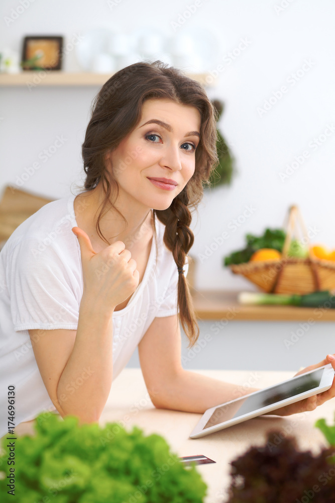 Young woman looking for a new recipe for cooking in a kitchen. Housewife is making online shopping by tablet computer and credit card Young woman looking for a new recipe for cooking in a kitchen. Housewife is making online shopping by tablet computer and credit card