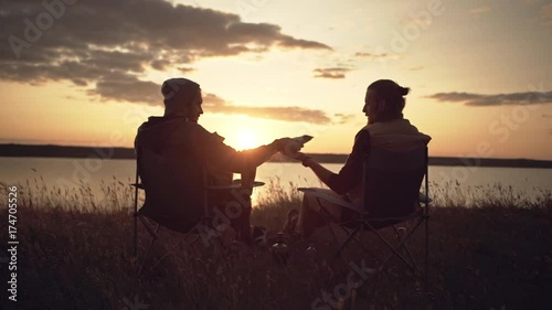 Two men friends camping near a river during sunset, passing food from a parcel. Slow motion.