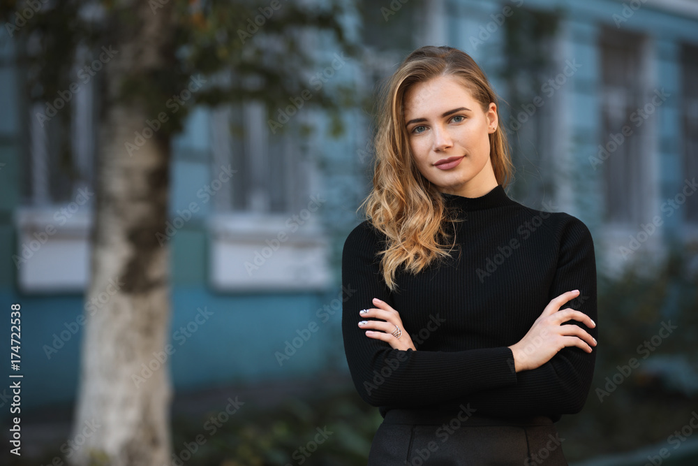 Portrait of young woman student against blue university building background, arms crossed Portrait of young woman student against blue university building background, arms crossed
