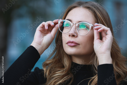 Myopia, close-up portrait of young woman student in eyeglasses for good vision looking up, blue building background photo