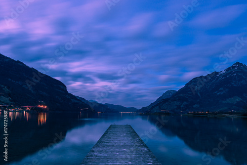 Wooden pier. Night landscape. Royal blue. The mountain range, the light of lanterns and lamps are reflected in the lake.