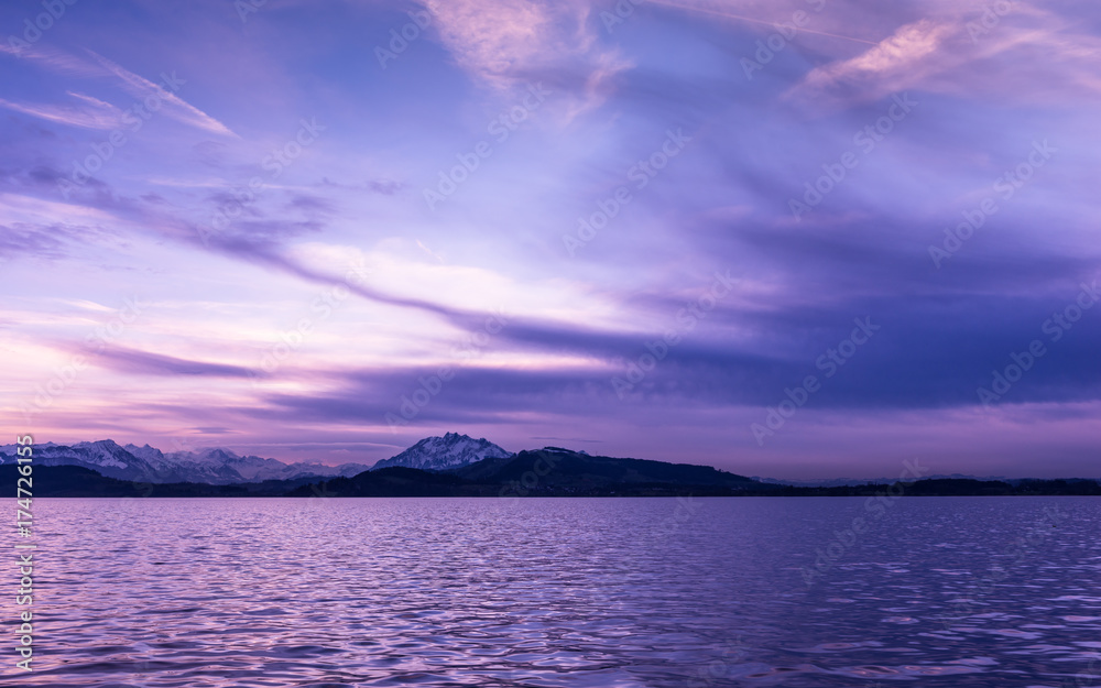 Luxury sunset in lilac over lake Zug. Switzerland. In the background silhouettes of the top of mountains in snow.