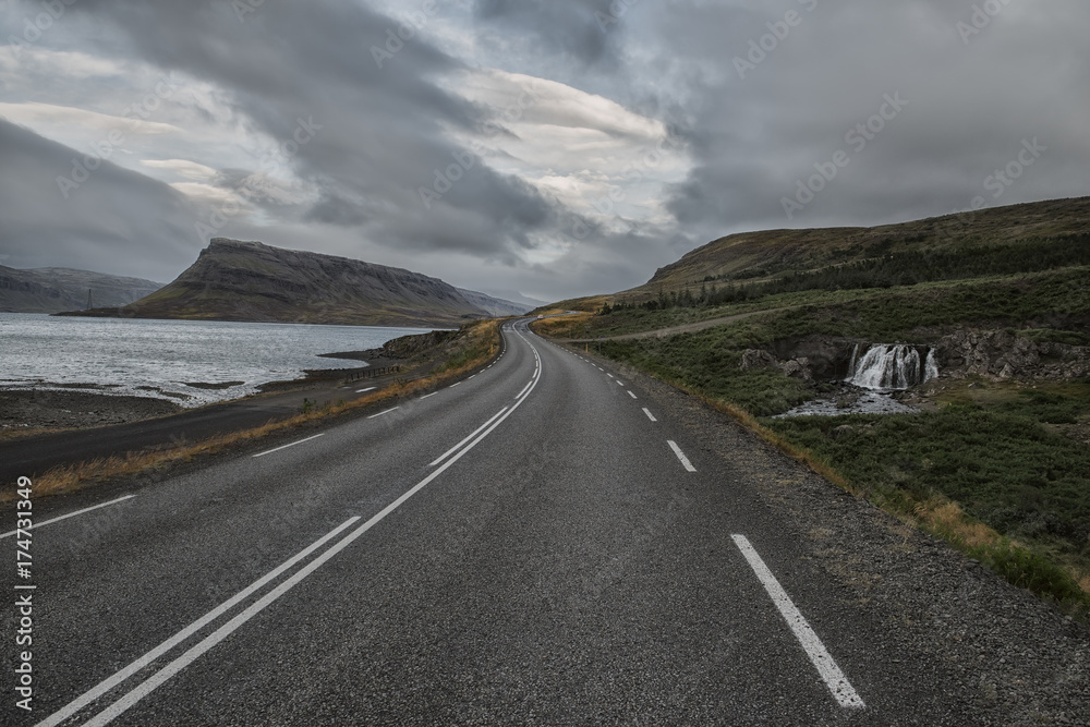 Fototapeta premium a deserted asphalt road running away into the hills, along the road a waterfall. Iceland. The spirit of travel and adventure. 