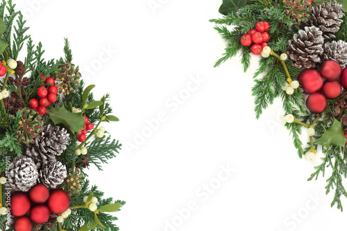 Christmas decorative border with red bauble decorations, holly, ivy, mistletoe, cedar and juniper leaf sprigs and pine cones on white background. 