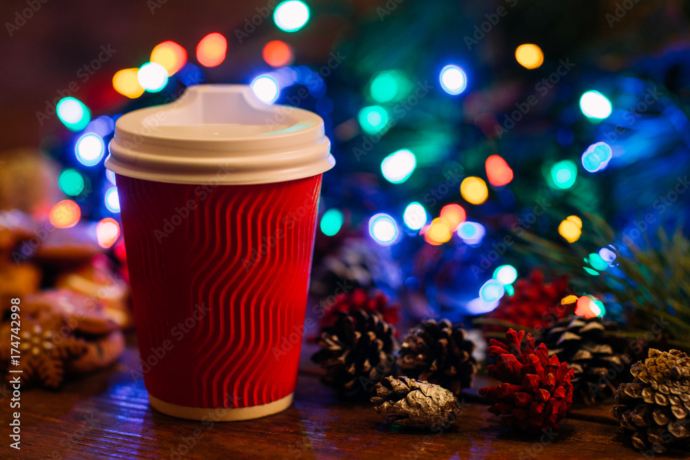 Delicious Christmas with take away latte. Close up cup of warm energy drink and strobila decoration on festive fairy lights background. Cozy xmas evening in cafe and fairs, happy holiday concept