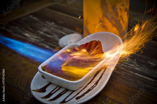Obraz na plátně Close-up of crème brûlée being torched, flames caramelizing sugar atop creamy dessert, capturing the fiery cooking process