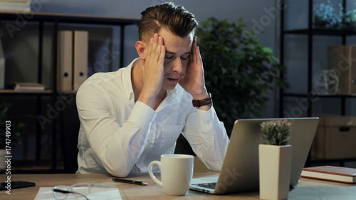 Portrait of tired business man in casual shirt working in the laptop in the stylish office, feeling very exausted. photo