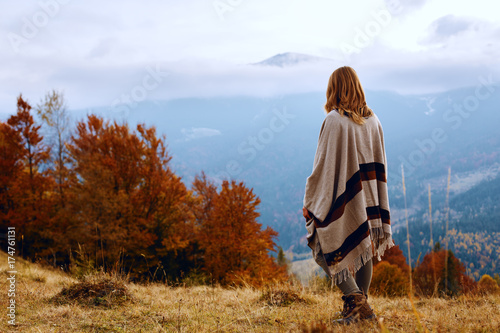 Hiker girl wrapping in warm poncho outdoor. Young woman hiking at mountain peak