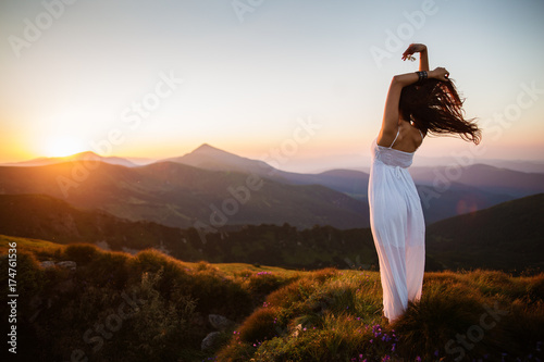 Hiker woman standing with hands up achieving the top. Girl welcomes a sun. Conceptual design. Successful woman hiker open arms on sunrise mountain top. Girl in long white dress in the mountains.