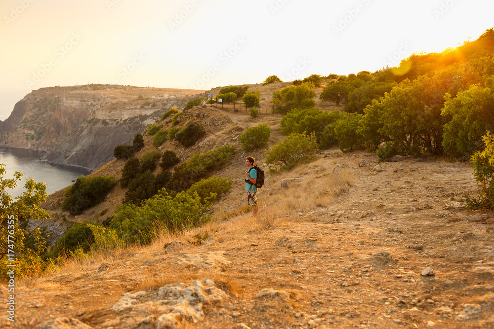 Fototapeta premium Among the fields and the sea, a tourist with a backpack enjoys the scenery at sunset and feels freedom, the view of the mountains can be seen from afar, the sun shines with bright rays.