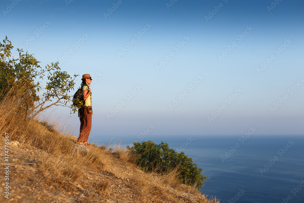 Traveling woman with backpack and hat looking at sea at sunny day. Adventure, travel people concept.
