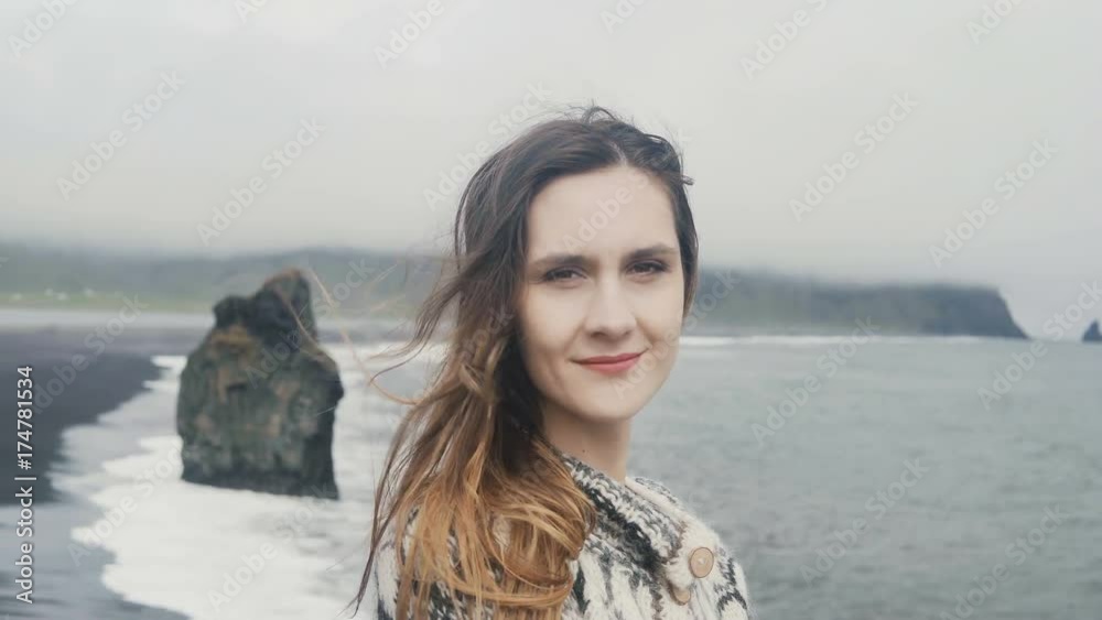 Portrait of young smiling woman standing on the black beach, troll toes on the background in Iceland, looking at camera Portrait of young smiling woman standing on the black beach, troll toes on the background in Iceland, looking at camera