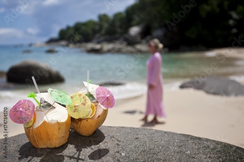 Two decorated coconuts filled with drinks standing on a granite rock, in the back a woman in a pink tunic, island Mahe, Seychelles, Indian Ocean, Africa