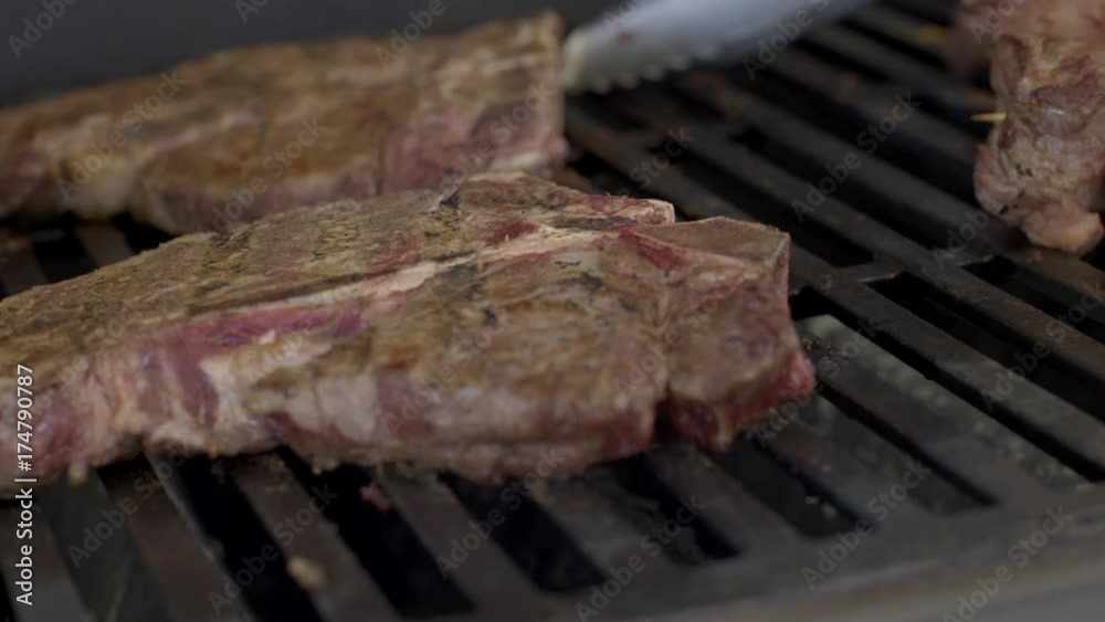Dolly move across T-bone steaks cooking on gas BBQ grill. Macro shot as tongs flip the meat, real time 4K. Dolly move across T-bone steaks cooking on gas BBQ grill. Macro shot as tongs flip the meat, real time 4K.