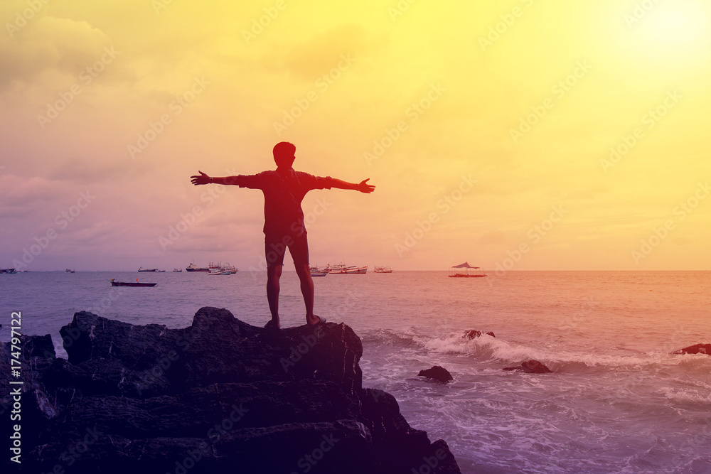 Silhouette hipster man standing raising hand on the rocks at the sea