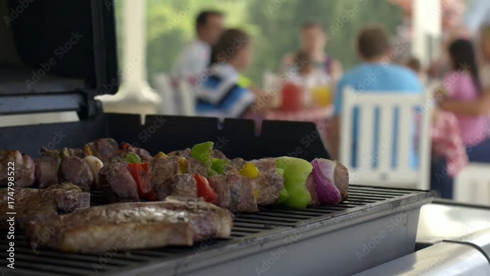 Dolly move around kebabs and steaks cooking on BBQ grill with family and friends soft focus, sitting at table in background.  Slow motion 4K recorded at 60fps.