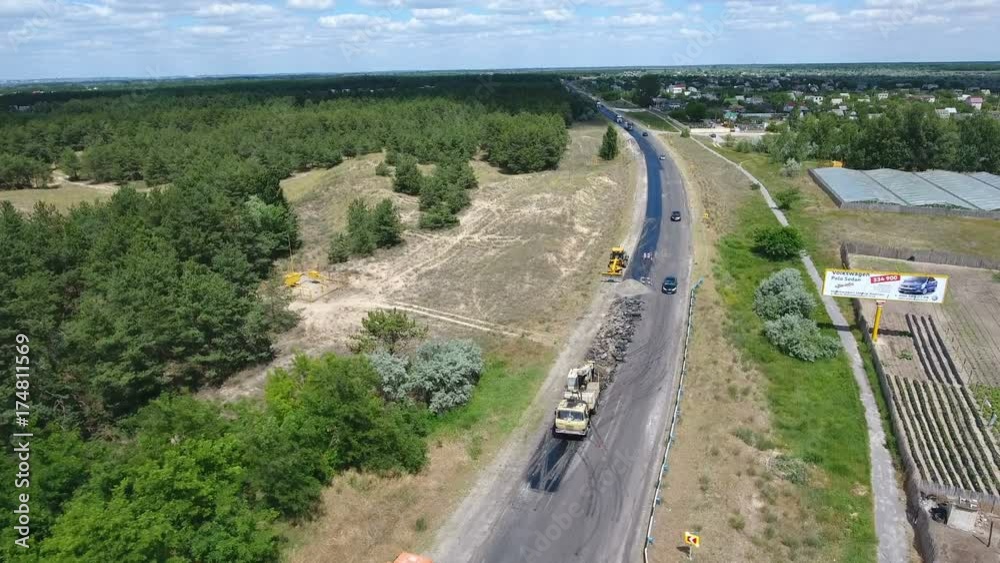 Impressive bird`s eye view of a road repairing and blacktopping with a steamroller, trucks and other vehicles standing nearby on a sunny day in summer. The greenary along the road looks fine. Impressive bird`s eye view of a road repairing and blacktopping with a steamroller, trucks and other vehicles standing nearby on a sunny day in summer. The greenary along the road looks fine.