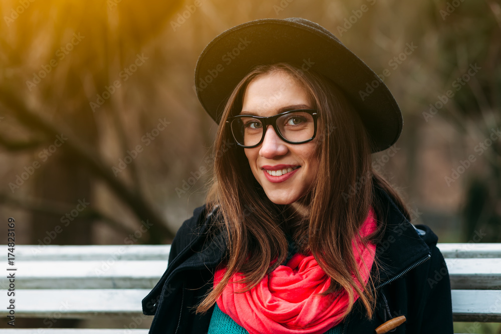 Close up portrait of beautiful smiling girl in hat and glasses sitting on bench in sunny autumn park