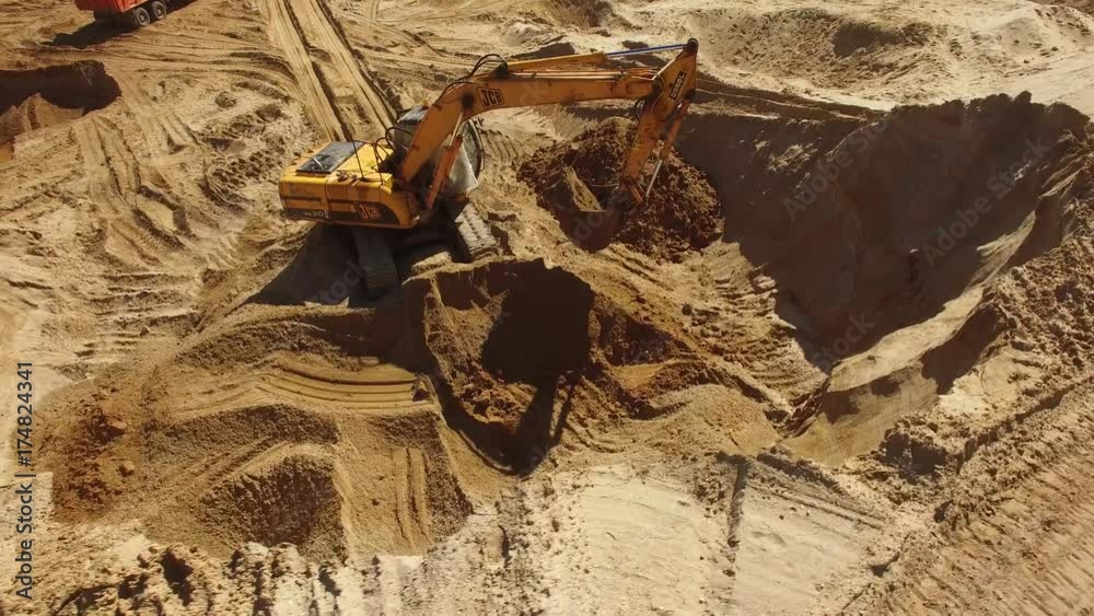 Excavator digging works in a quarry. Orange sand soil rock heavy industry heavy equipment. Aerial drone action flight around. Sunny day.
