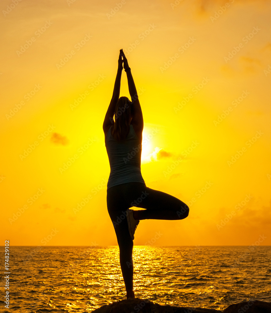 A young woman is practicing yoga on the rocks by the sea against the background of the sun

