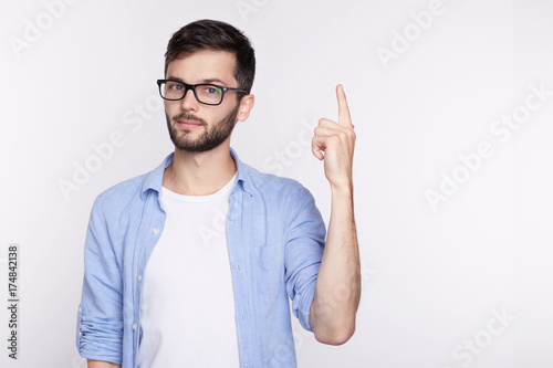 Waist up shot of joyful guy wearing blue long sleeve shirt and glasses looking up, pointing finger at copy space above his head. Bearded young man indicating something on blank studio wall with hand.