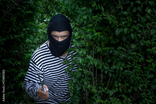 Foto A terrorist or robber is standing behind the bush with knife and  looking at cam