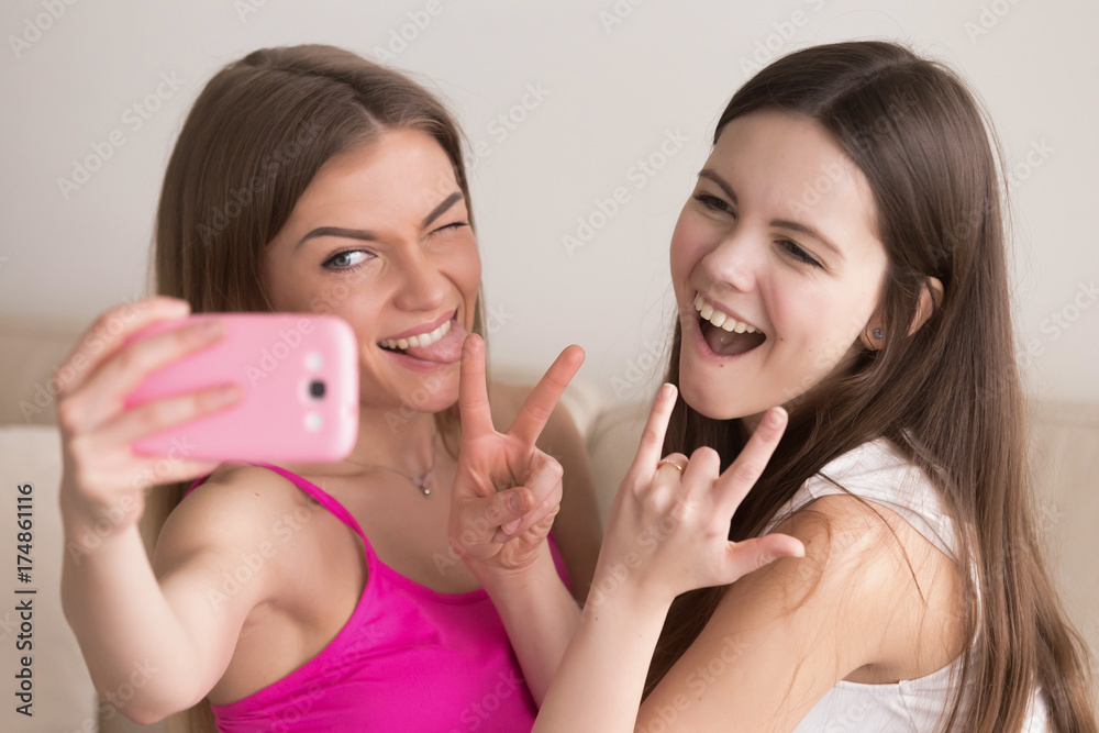 Two girlfriends taking selfie with smartphone, making funny faces, showing  peace and rock on signs. Smiling girls make memories with photos together.  Best friends take pictures for social media site. Stock Photo |