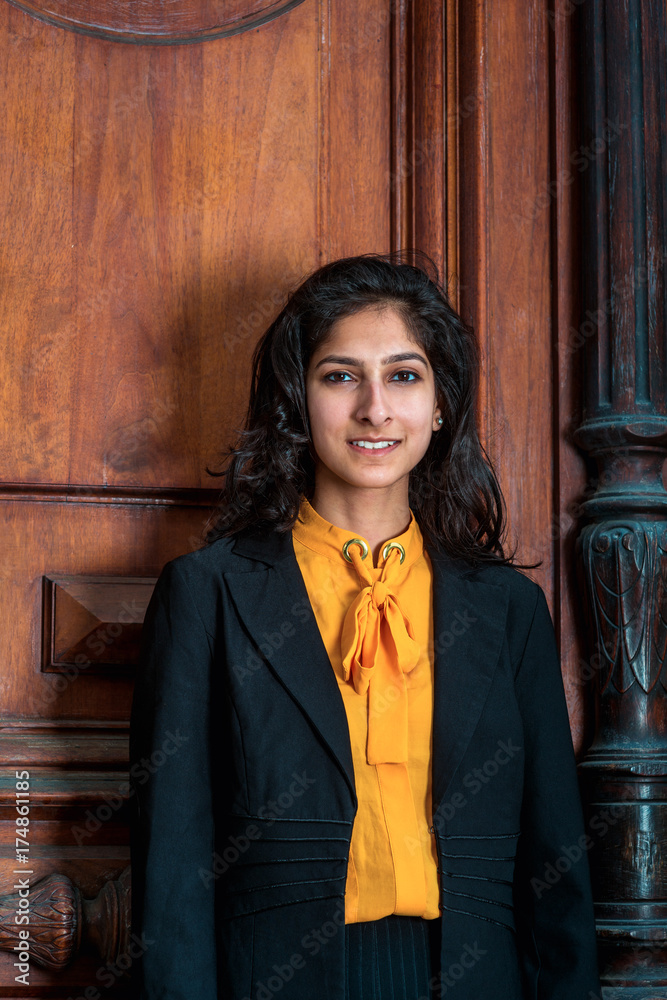 Modern East Indian American Woman with long curly hair in New York. Wearing  black blazer, orange under shirt, a college student standing by vintage  style office doorway, smile, looking at you.. Stock