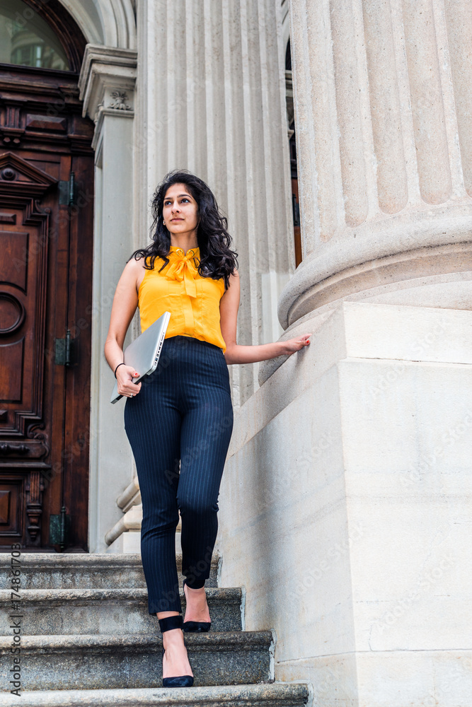 East Indian American college student studying in New York, wearing sleeveless orange shirt, striped pants, high heels, carrying laptop computer, walking down stairs. Filtered look with dark blue tint.