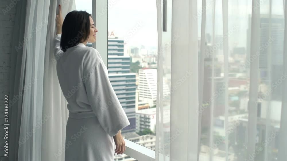 Woman in bathrobe unveil curtains and admire view from window at home
Woman in bathrobe unveil curtains and admire view from window at home