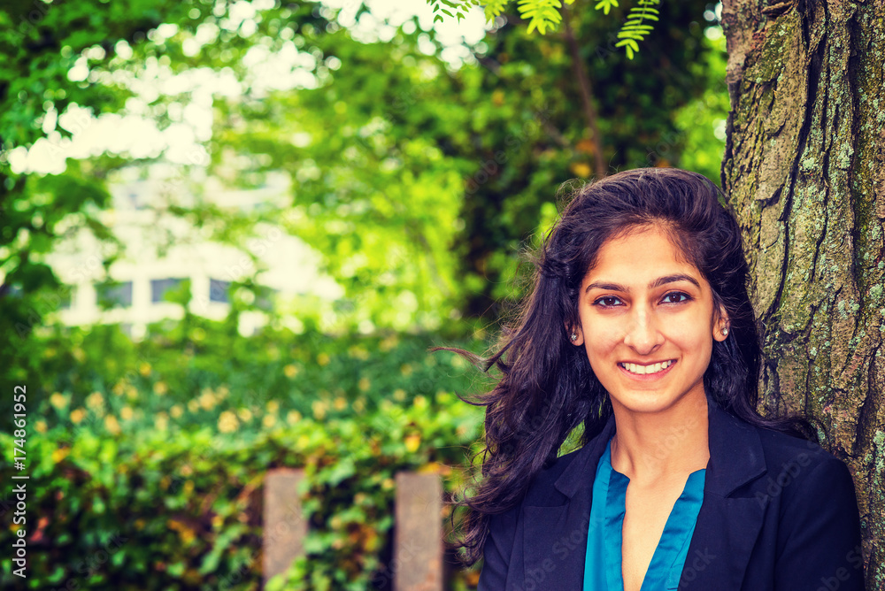 Happy East Indian American college student studying in New York, wearing black blazer, standing against tree trunk on green campus, smiling, waiting for classmates, friends. Instagram filtered effect. Happy East Indian American college student studying in New York, wearing black blazer, standing against tree trunk on green campus, smiling, waiting for classmates, friends. Instagram filtered effect.