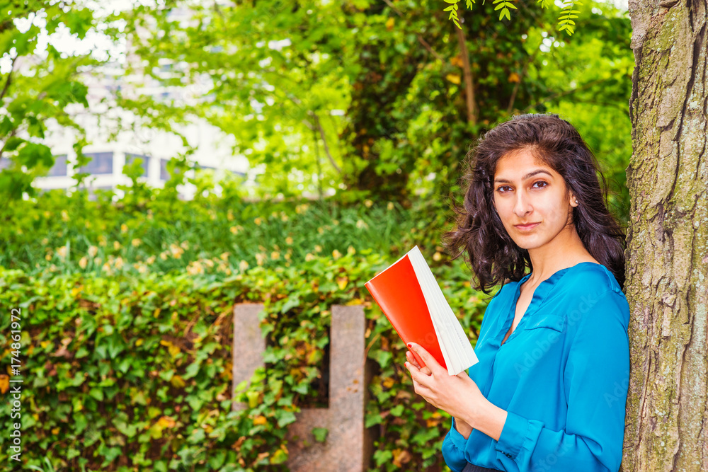 Wearing long sleeve blue shirt, hands holding a red book, a young East Indian American college student standing by tree on campus in New York, reading, enjoying outdoor fresh air in spring day.. Wearing long sleeve blue shirt, hands holding a red book, a young East Indian American college student standing by tree on campus in New York, reading, enjoying outdoor fresh air in spring day..