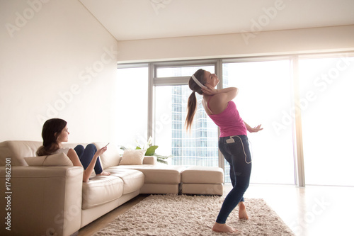 Young happy attractive girl in stylish white headphones listening and dancing to favorite music on smartphone in sunny living room at home. Joyful girlfriend relaxing on sofa with cellphone in hand.