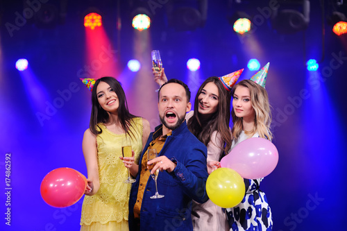 a young man in the company of three pretty girls is celebrating the event with champagne glasses in hand, balloons and hats for a holiday on the head.
