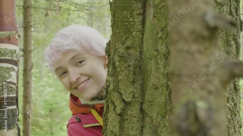 Young attractive smiling woman looking behind the tree. Blonde girl check and looks from behind a tree In the autumn forest photo