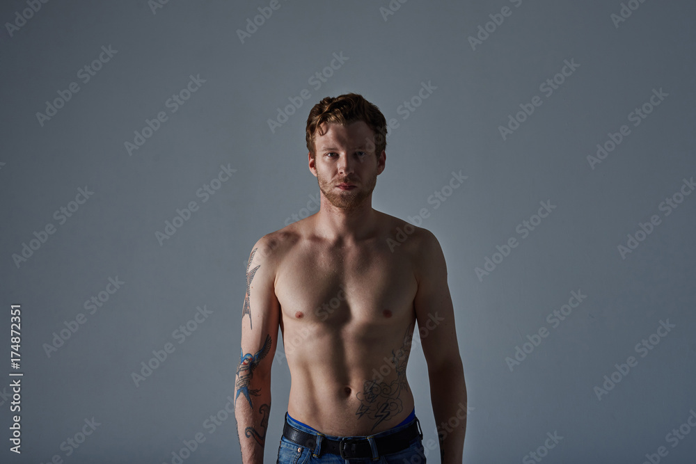 Waist up portrait of attractive adult confident man with beard and muscular abdominal posing shirtless against blank studio wall background with copy space for your text or promotional content