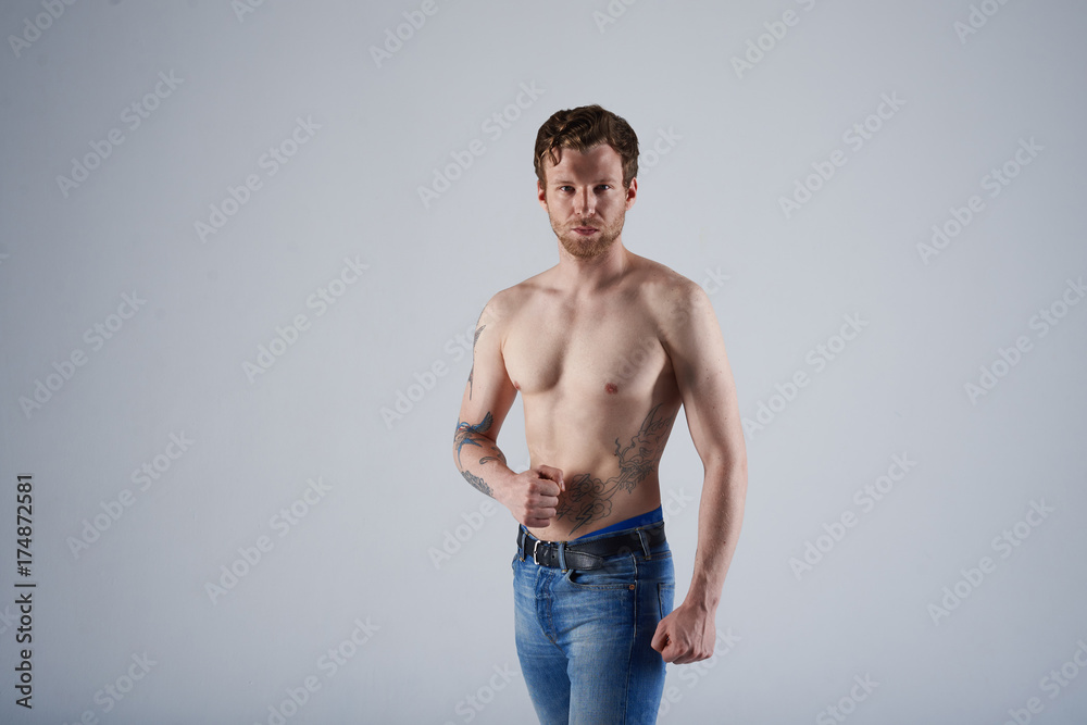 Bearded young sportsman with nude torso posing at blank grey wall, tensing muscles, showing muscular arms at camera, having serious look. Confident macho man with beard and tattoos modeling