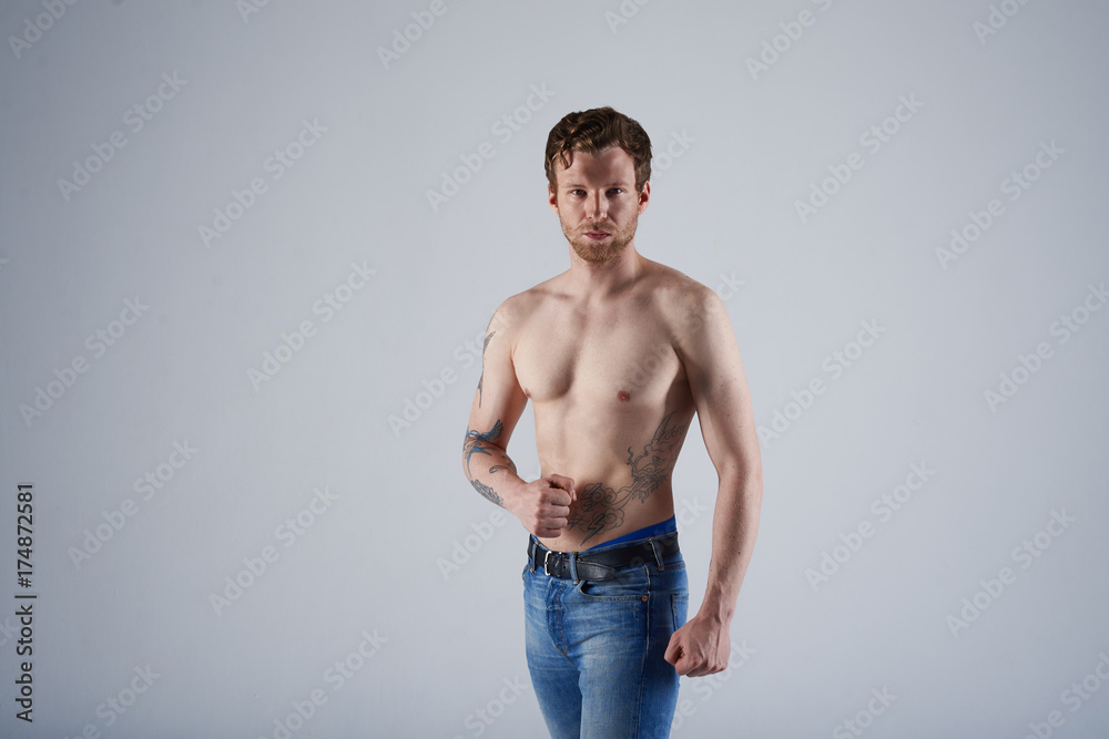 Bearded young sportsman with nude torso posing at blank grey wall, tensing muscles, showing muscular arms at camera, having serious look. Confident macho man with beard and tattoos modeling Bearded young sportsman with nude torso posing at blank grey wall, tensing muscles, showing muscular arms at camera, having serious look. Confident macho man with beard and tattoos modeling