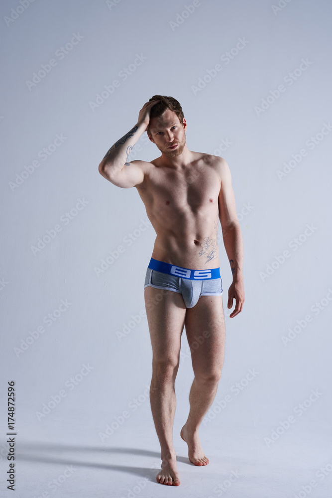 Vertical isolated shot of attractive sexy man standing in white studio wearing no clothes except blue and white pants. Bearded guy advertising high fashion underwear, adjusting his ginger hair