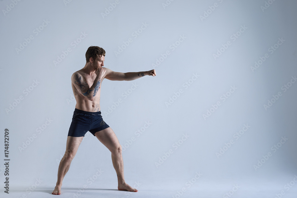 Full length shot of muscular athletic young Caucasian kickboxer training in studio, punching air, preparing for fight, wearing black shorts and no shoes. People, sports, boxing and fighting
