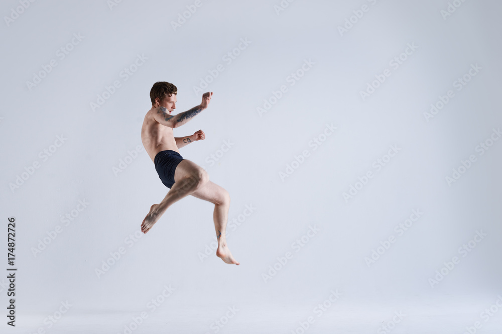 Freeze action shot of flying shirtless and barefooted young male boxer wearing trunks jumping high against blank grey studio wall background with copy space for your advertising information