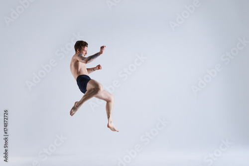 Freeze action shot of flying shirtless and barefooted young male boxer wearing trunks jumping high against blank grey studio wall background with copy space for your advertising information