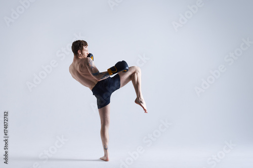 Full lenght shot of shirtless and barefooted young Caucasian male fighter raising one leg, ready to kick invisible enemy. Boxing, kickboxing, martial arts, sports, power and strenght concept