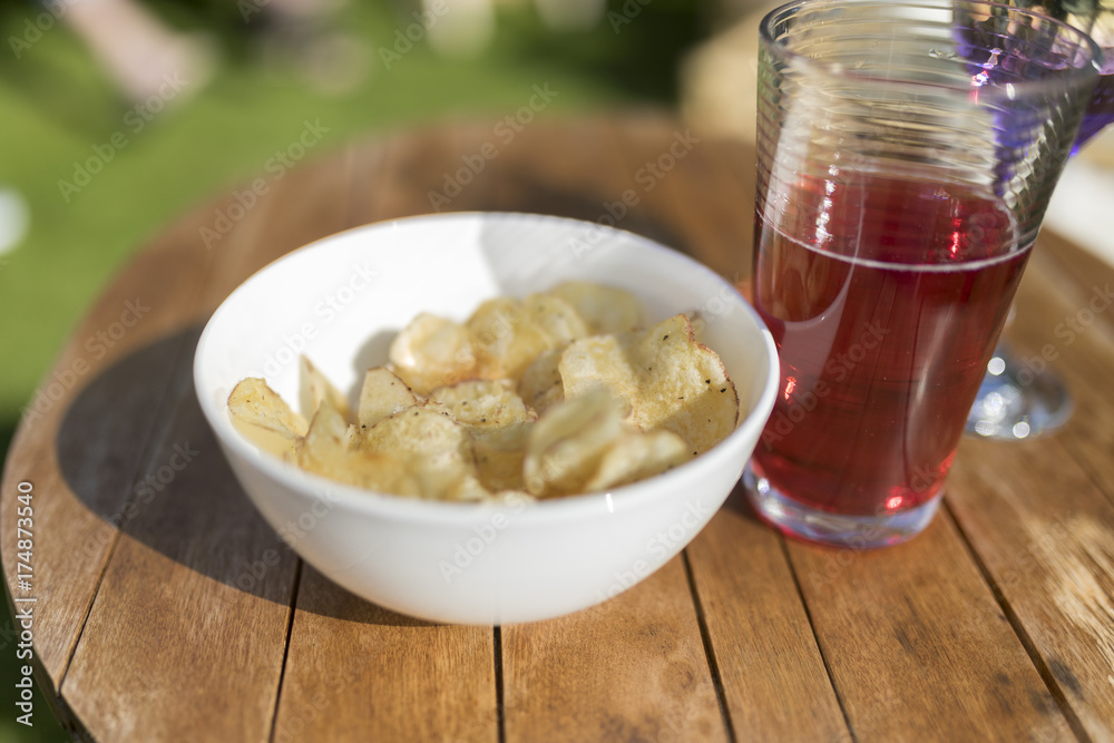 Single white bowl of crisps and a glass of juice outside on a wooden table in summer Single white bowl of crisps and a glass of juice outside on a wooden table in summer